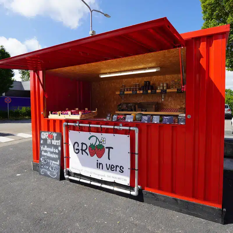 Een felrode metalen container omgebouwd tot een marktkraam, met een open luik dat als afdak dient. De kraam verkoopt verse producten zoals Hollandse aardbeien, uitgestald in bakjes op een houten toonbank. Aan de voorkant hangt een bord met de tekst 'Groot in vers' en een aardbeienlogo. Daarnaast staat een krijtbord met productprijzen en teksten. De achtergrond toont een weg en wat groen.