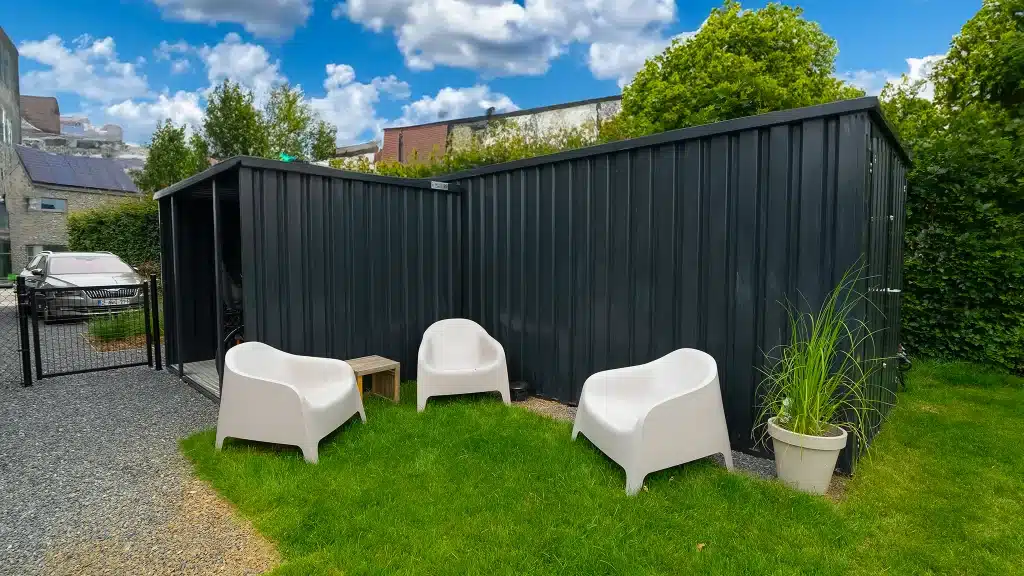 Een moderne, zwarte metalen overkapping en schutting vormen een strakke afscheiding in een tuin met groen gras en grind. Drie witte kunststof stoelen staan in een halve cirkel rond een klein, vierkant houten tafeltje. Rechts van de stoelen staat een witte bloempot met een hoge groene plant. Op de achtergrond zijn bomen en huizen zichtbaar onder een blauwe lucht met wolken.