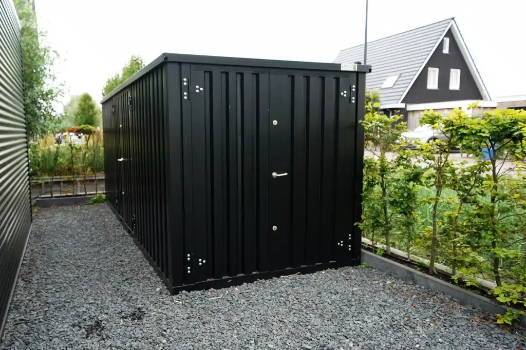 Een grote zwarte metalen opslagcontainer met dubbele deuren en zilverkleurige scharnieren, geplaatst op een ondergrond van grijs grind. De container staat naast een moderne woning met een donker zadeldak en wordt omgeven door een rij jonge bomen en groene struiken.