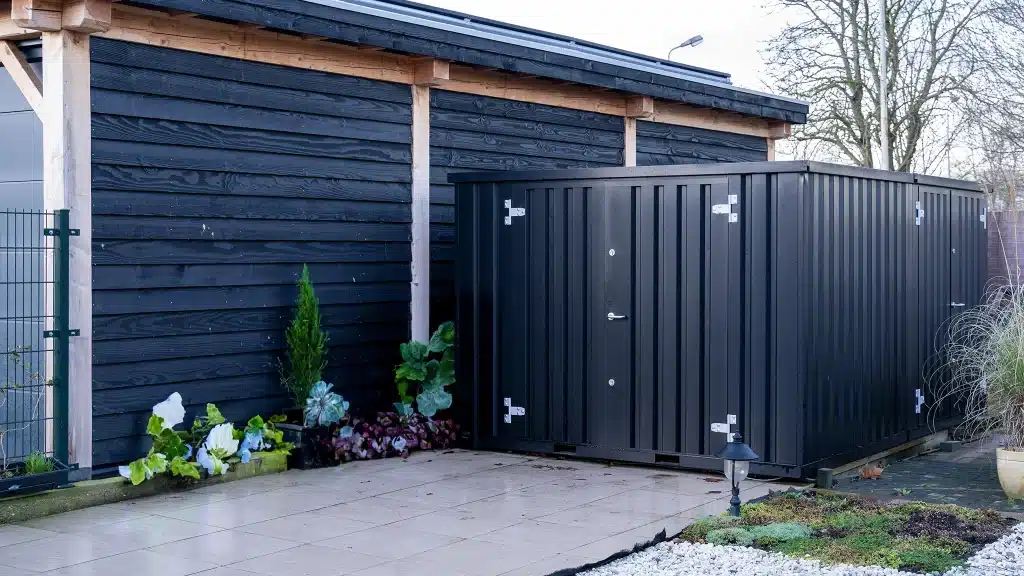 Een zwarte metalen opslagcontainer met zilverkleurige scharnieren en een deur, geplaatst op een betegeld terras naast een zwarte houten schuur met houten steunbalken. De container wordt omringd door sierplanten en grind, met kale bomen zichtbaar op de achtergrond onder een heldere hemel