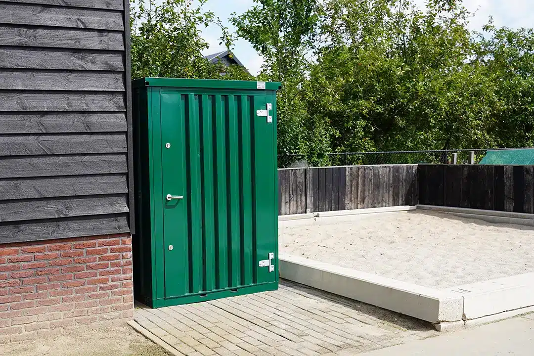 Een groene metalen opslagcontainer met een gesloten deur en zilverkleurige scharnieren, geplaatst tegen de gevel van een gebouw met zwarte houten panelen. De container staat op een houten terras, met een verhoogd betegeld gebied en groene bomen op de achtergrond onder een heldere blauwe lucht.
