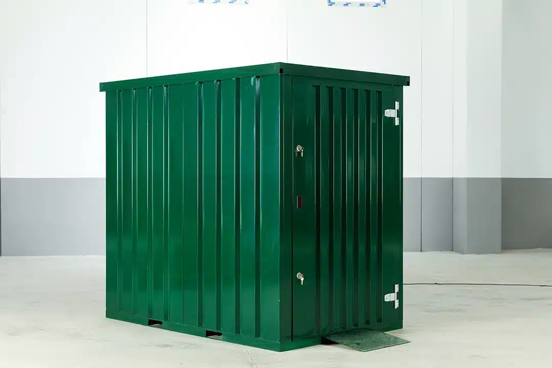 Een donkergroene, metalen opbergcontainer staat op een schone, lichtgekleurde vloer in een minimalistische binnenruimte. De container heeft een stevige structuur met verticale ribbels, een deur met een slot en scharnieren aan de rechterkant. Een kleine mat ligt voor de deur, en de achtergrond bestaat uit een gladde witte muur met grijze accenten.
