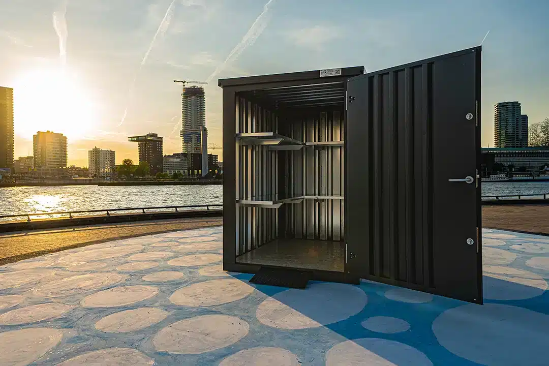 Een open zwarte metalen opslagcontainer met zichtbare metalen schappen aan de binnenkant. De container staat op een kleurrijke bestrating met blauwe en witte cirkels, vlak bij een kade met uitzicht op een rivier. Op de achtergrond is een skyline met hoge gebouwen zichtbaar tijdens een sfeervolle zonsondergang.