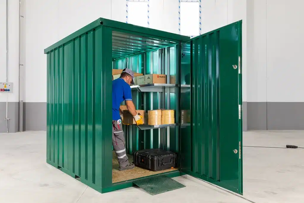 Een groene metalen opslagcontainer met open deuren, waarin een man in werkkleding dozen aan het plaatsen is op metalen schappen. De container staat in een grote lichte binnenruimte met witte muren, een grijze onderrand en een betonnen vloer. Er ligt een kleine oprit bij de ingang van de container.