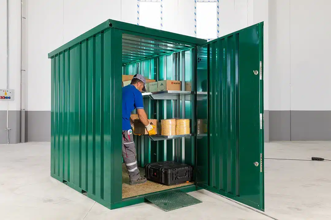 Een groene metalen opslagcontainer met open deuren, waarin een man in werkkleding dozen aan het plaatsen is op metalen schappen. De container staat in een grote lichte binnenruimte met witte muren, een grijze onderrand en een betonnen vloer. Er ligt een kleine oprit bij de ingang van de container.
