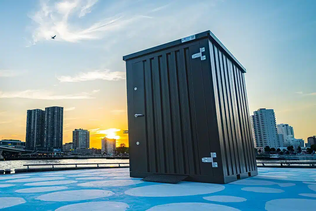 Een zwarte metalen opslagcontainer met zilverkleurige scharnieren, geplaatst op een kleurrijk terras met blauwe en witte patronen. Op de achtergrond is een rivier te zien met hoge gebouwen en een sfeervolle zonsondergang die de lucht oranje kleurt.
