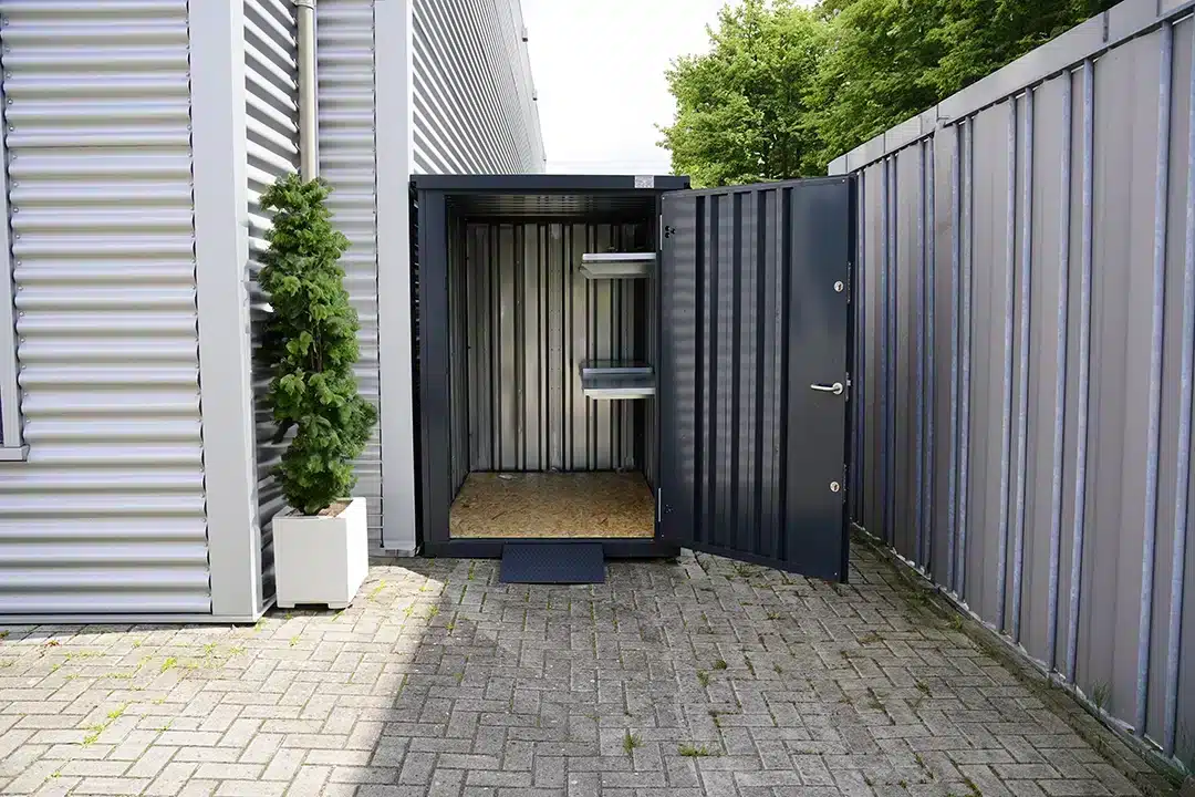 Een open antracietgrijze metalen opslagcontainer met zichtbare binnenkant, ingericht met metalen schappen en een houten vloer. De container staat op een betegeld terrein naast een gebouw met een geribbelde metalen gevel en een hekwerk. Een kleine conifeer in een witte plantenbak staat links van de container, met bomen zichtbaar in de achtergrond.