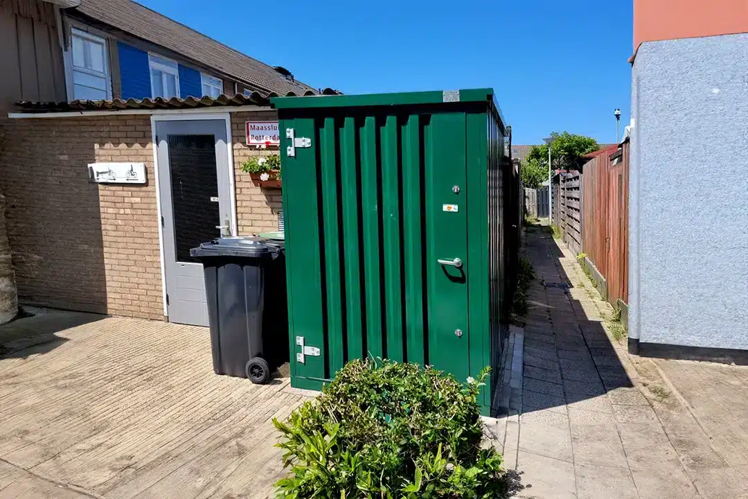 Een groene metalen opslagcontainer met een gesloten deur, geplaatst op een betegeld pad naast een huis. Naast de container staan een zwarte afvalcontainer en een groene struik. Op de achtergrond zijn een bakstenen huis, een schutting en een blauwe lucht zichtbaar.