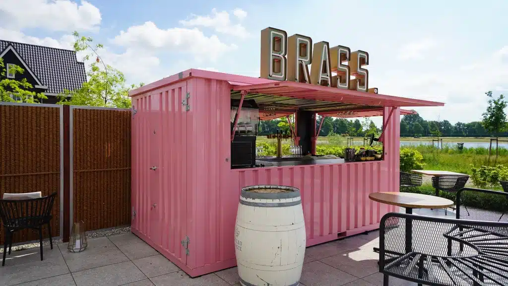 Een felroze metalen container omgebouwd tot een bar met het grote bord 'BRASS.' bovenop. De bar heeft open luiken, waar de toonbank met apparatuur zichtbaar is. De container staat op een betegeld terras met tafels, stoelen en een wit wijnvat als decoratief element. Op de achtergrond is een groen landschap zichtbaar met bomen, struiken en water onder een blauwe lucht.