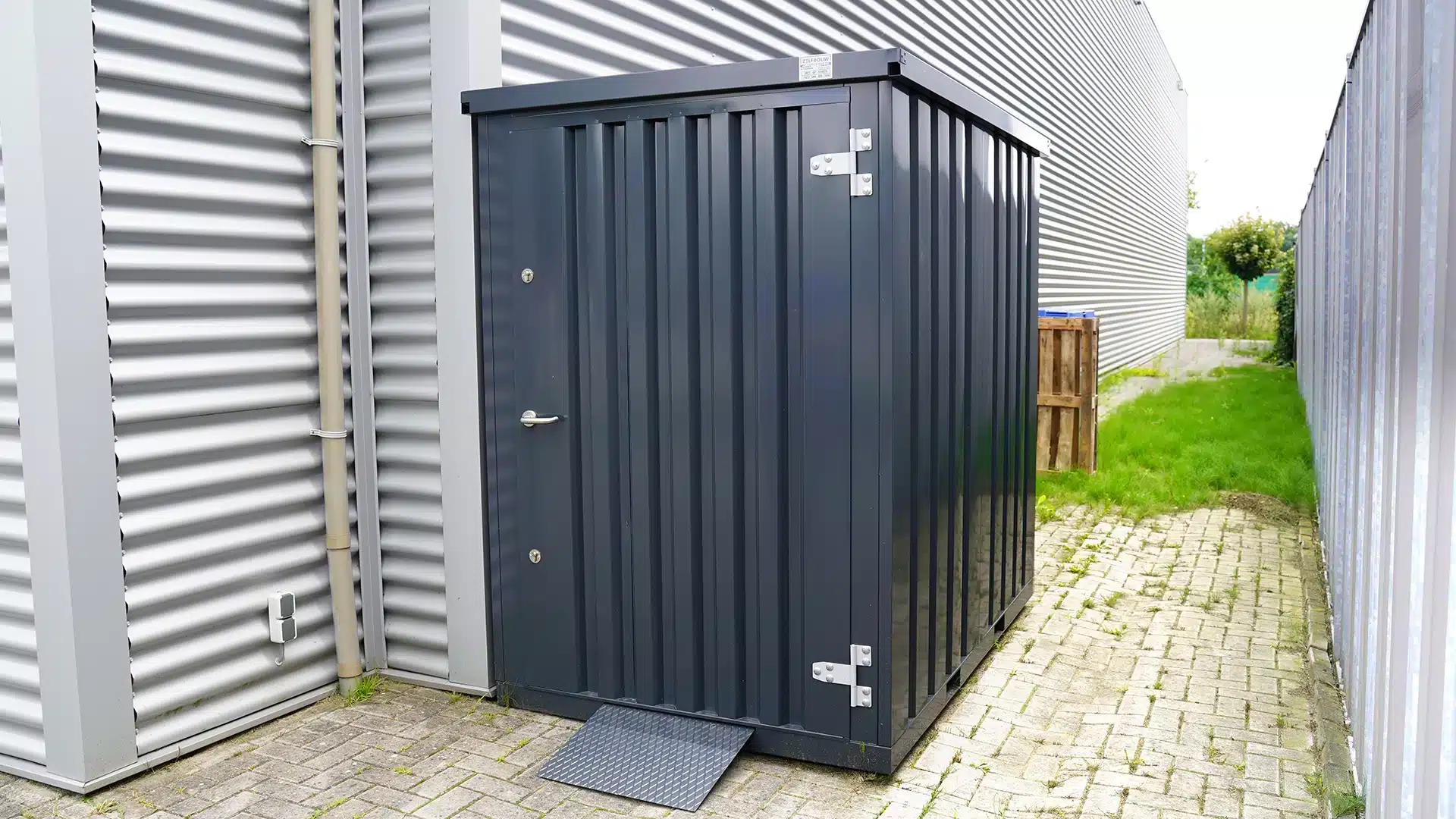 Donkergrijze demontabele opslagcontainer met enkele deur, geplaatst buiten tussen twee bedrijfspanden