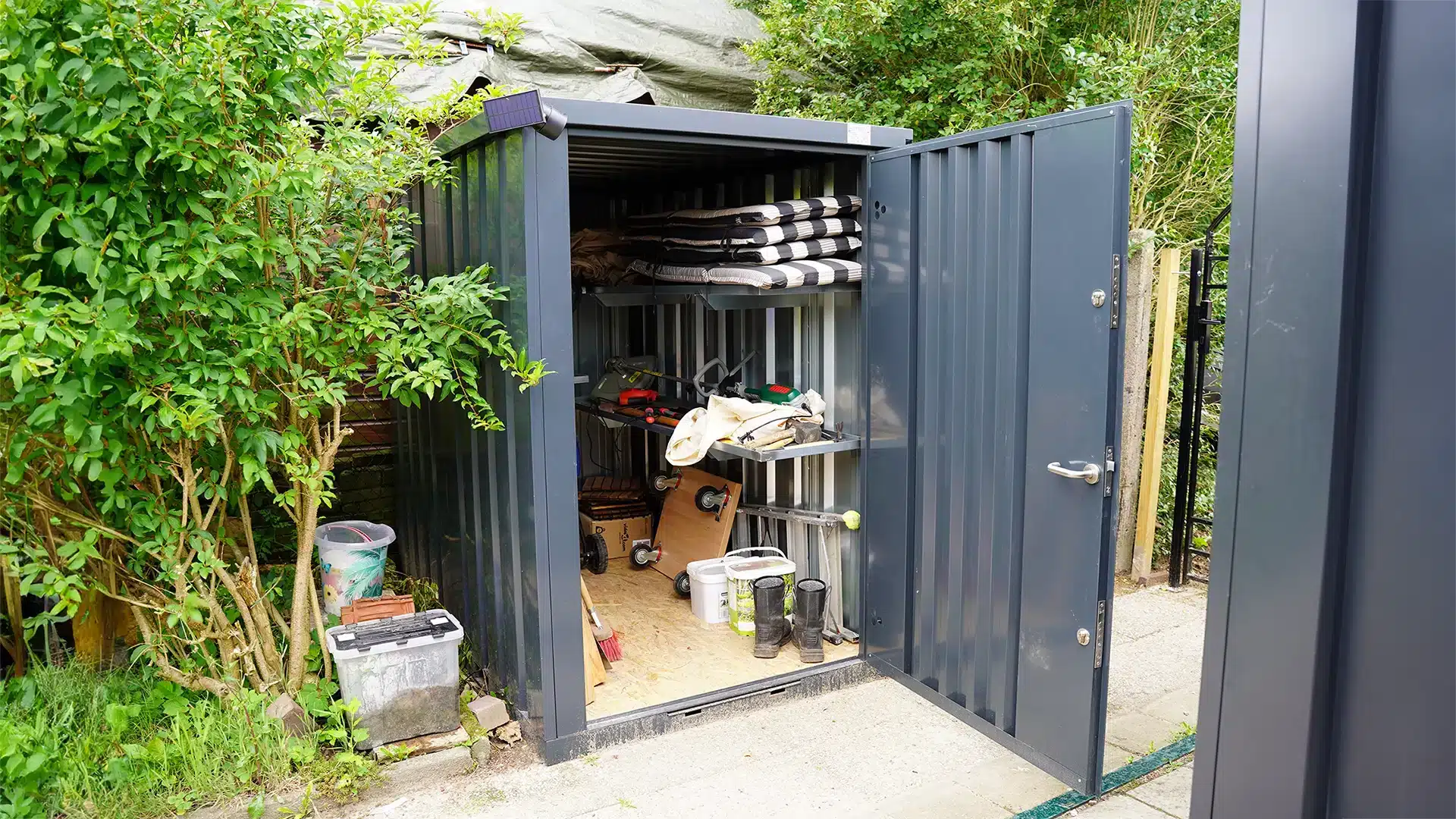Opengeslagen metalen opslagcontainer met gereedschap, laarzen en opbergsystemen tussen groen in een tuin