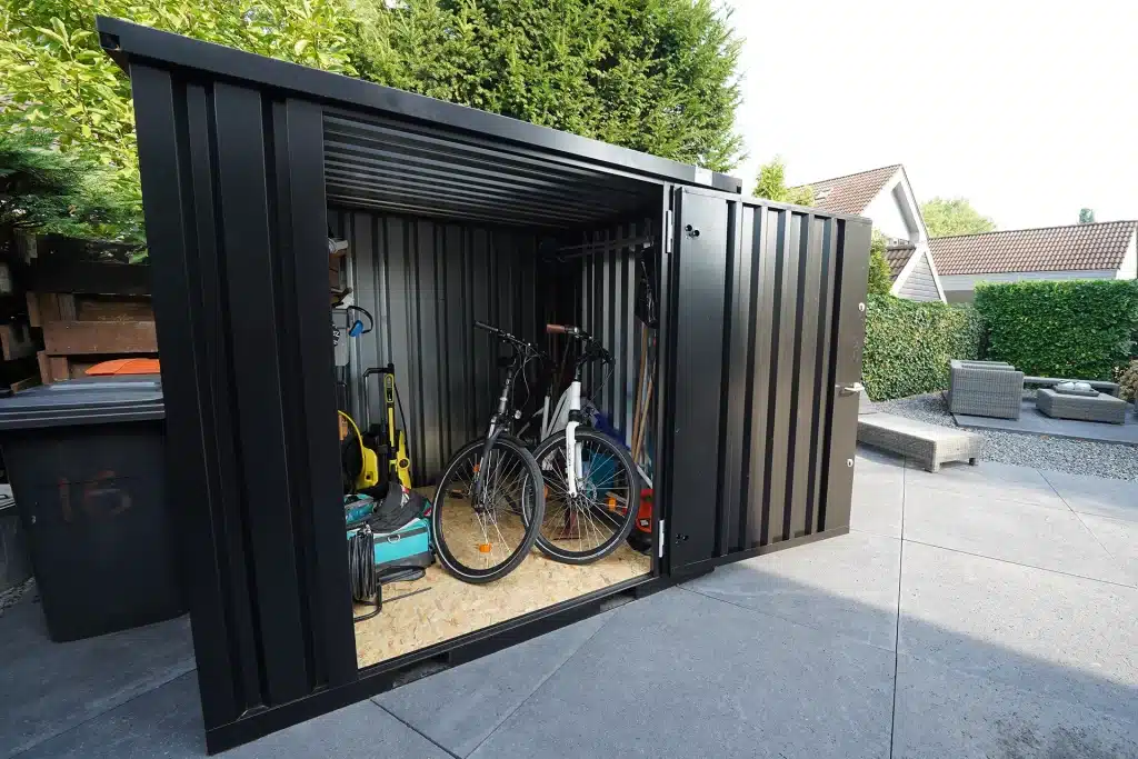 Een geopende zwarte metalen 10 ft opslagcontainer waarin meerdere fietsen, tuingereedschap en andere spullen zoals een hogedrukreiniger en een tuinslang zijn opgeborgen. De container staat op een betegeld terras in een tuin, omringd door groene heggen en bomen. Op de achtergrond zijn een grijze loungebank en een deel van een huis zichtbaar.