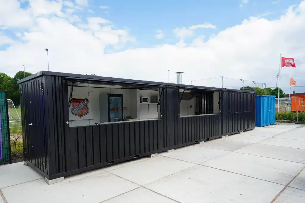 Een lange zwarte container met meerdere ramen en een deur, ingericht als kiosk of kantine. Binnen zijn een koelkast, magnetron en andere apparatuur zichtbaar. De container staat op een verhard terrein naast een sportveld, met hekken en vlaggenmasten op de achtergrond. Er wappert een rode vlag en de lucht is licht bewolkt.