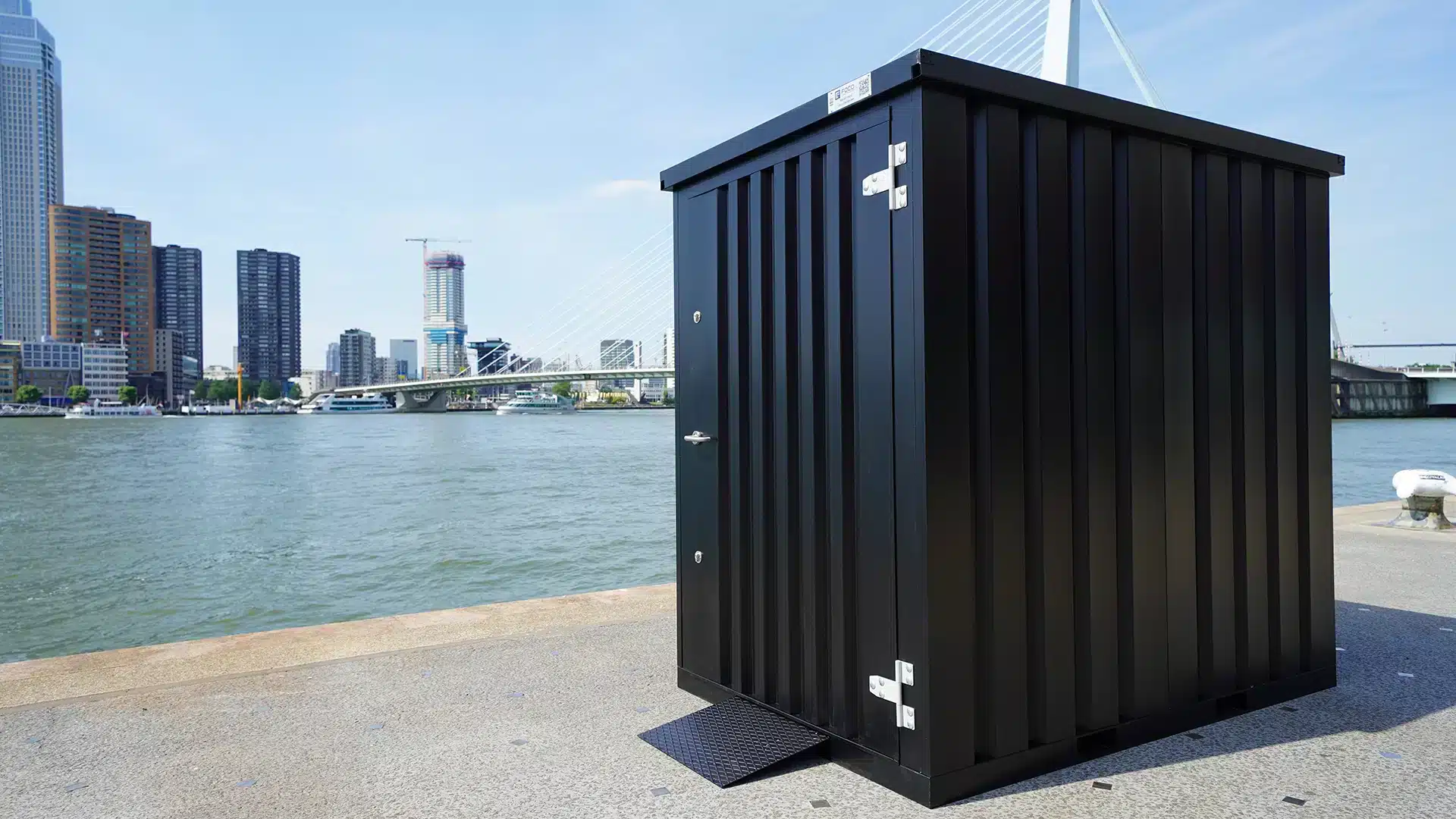 Zwarte stalen opslagcontainer met dubbele deur, geplaatst aan de kade in Rotterdam met uitzicht op de Erasmusbrug