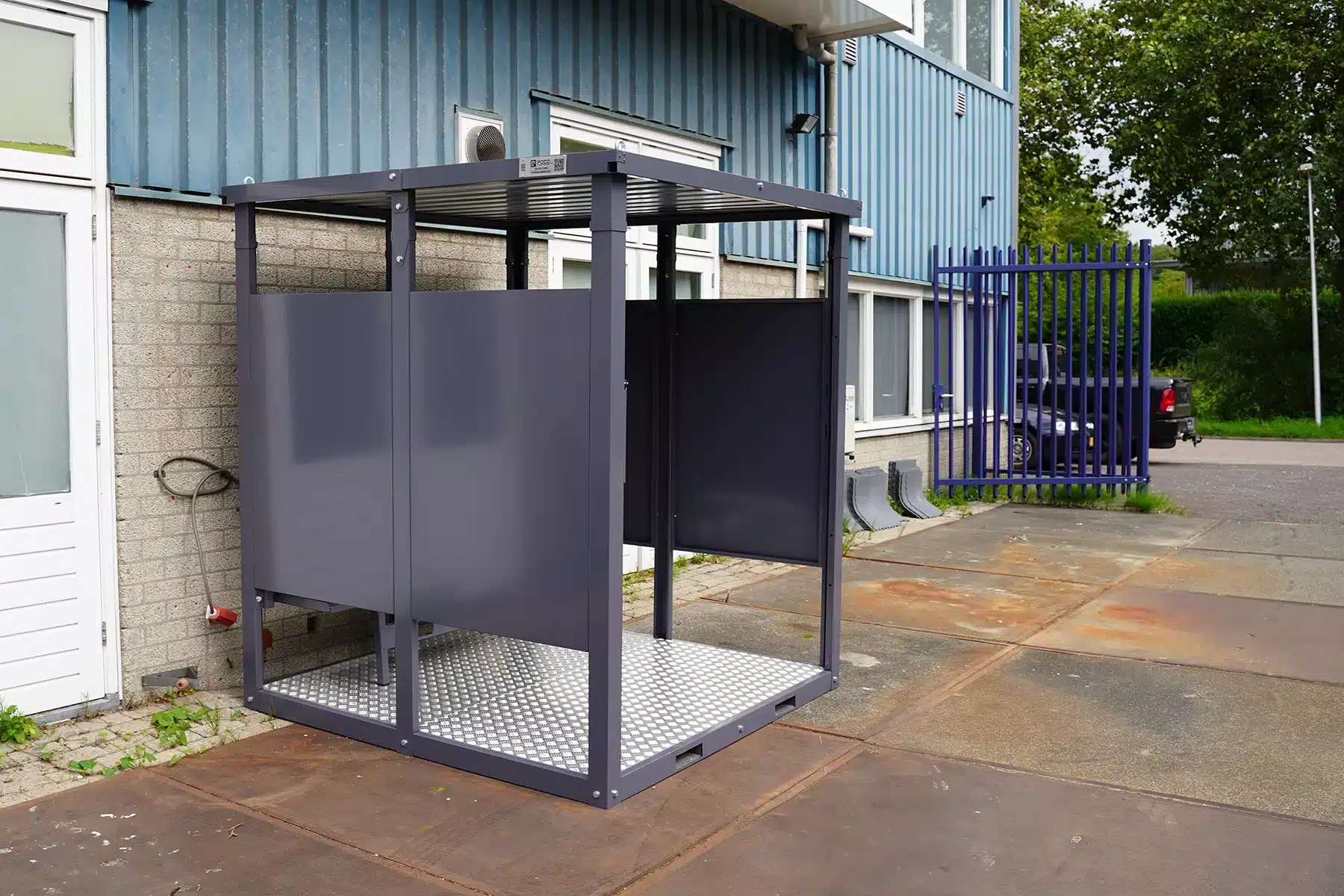 Een compacte donkergrijze metalen rookruimte met een stevig dak en verhoogde antislip metalen vloer. De structuur is gedeeltelijk open aan de voorzijde en heeft een ingebouwde zitbank. De rookruimte staat op een verhard industrieterrein naast een gebouw met blauwe gevelbekleding en een gesloten hek in de achtergrond.