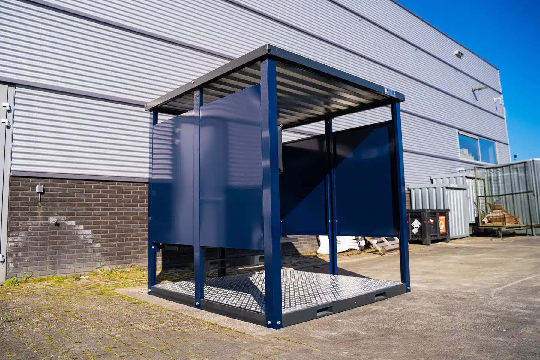 Een blauwe metalen overkapping met een gedeeltelijk open ontwerp en een golfplaten dak staat op een industrieterrein. Hij dient als rookcontainer. De structuur heeft vier stevige pilaren en een verhoogde metalen vloer met een antislip patroon. De achtergrond toont een groot industrieel gebouw met grijze en witte gevelpanelen en enkele ramen