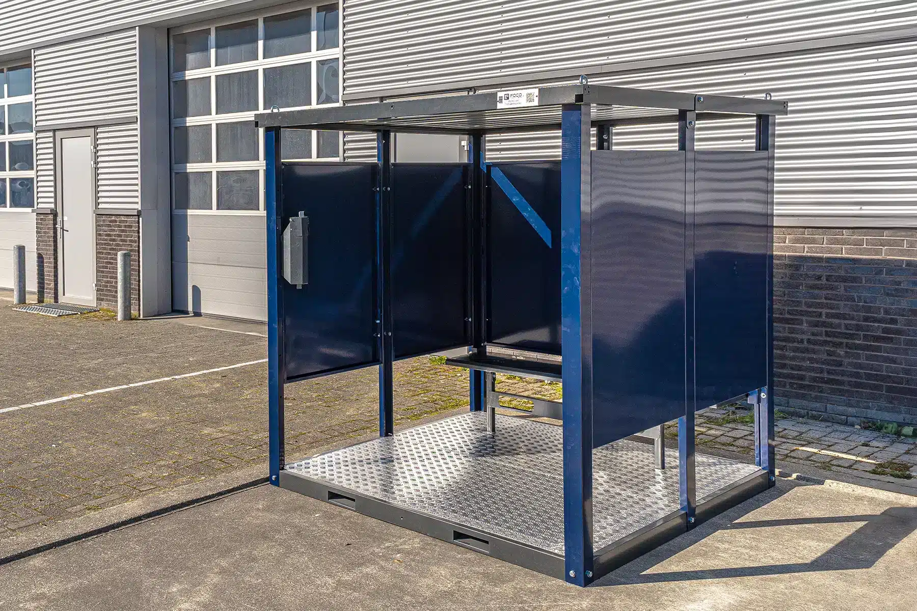 Een stevige blauwe metalen rookruimte met een gedeeltelijk open ontwerp, voorzien van een verhoogde metalen vloer met antislip patroon en een dak. De ruimte heeft een ingebouwde metalen bank. Op de achtergrond staat een groot industrieel gebouw met een grijze gevel, ramen en een garagepoort.