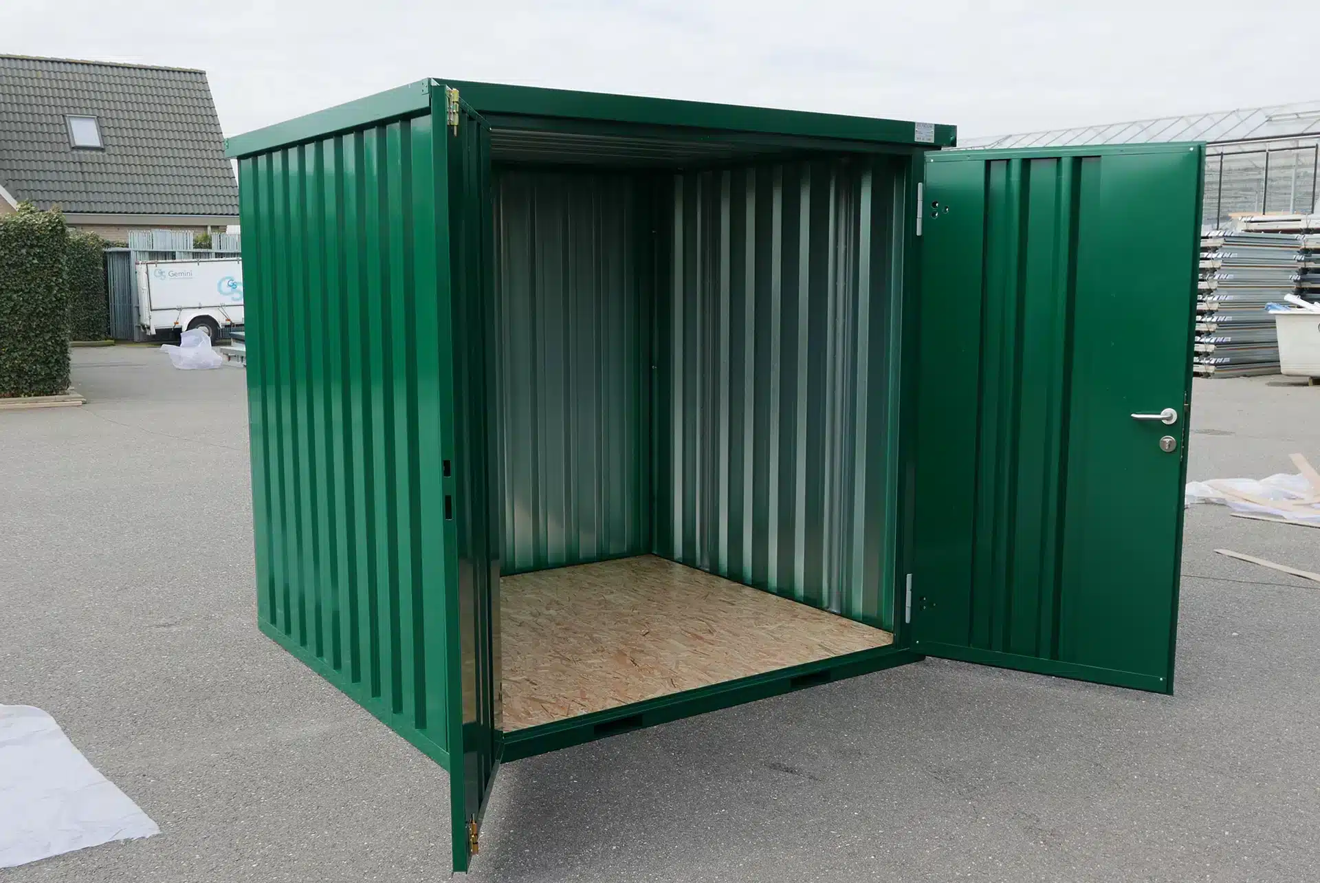 Een open groene metalen opslagcontainer met zicht op de binnenkant, voorzien van een houten vloer en metalen wanden. De container staat op een verhard terrein, met gebouwen, een heg en diverse objecten op de achtergrond onder een bewolkte hemel.