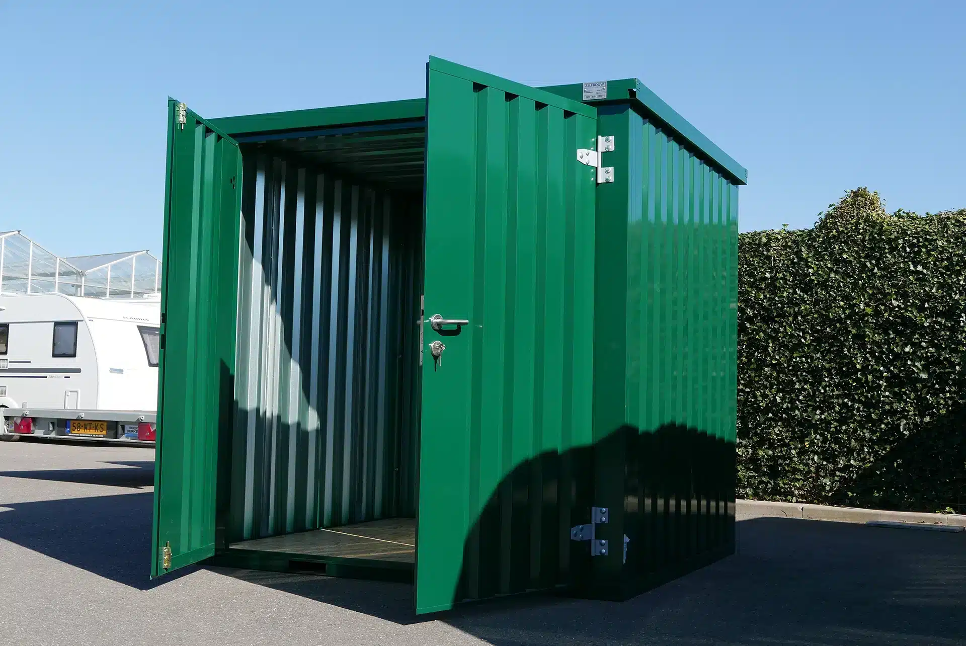 Een open groene metalen opslagcontainer met een zichtbare binnenkant, bestaande uit metalen wanden en een houten vloer. De container staat op een verharde ondergrond, met een heg en een camper zichtbaar op de achtergrond onder een heldere blauwe lucht.