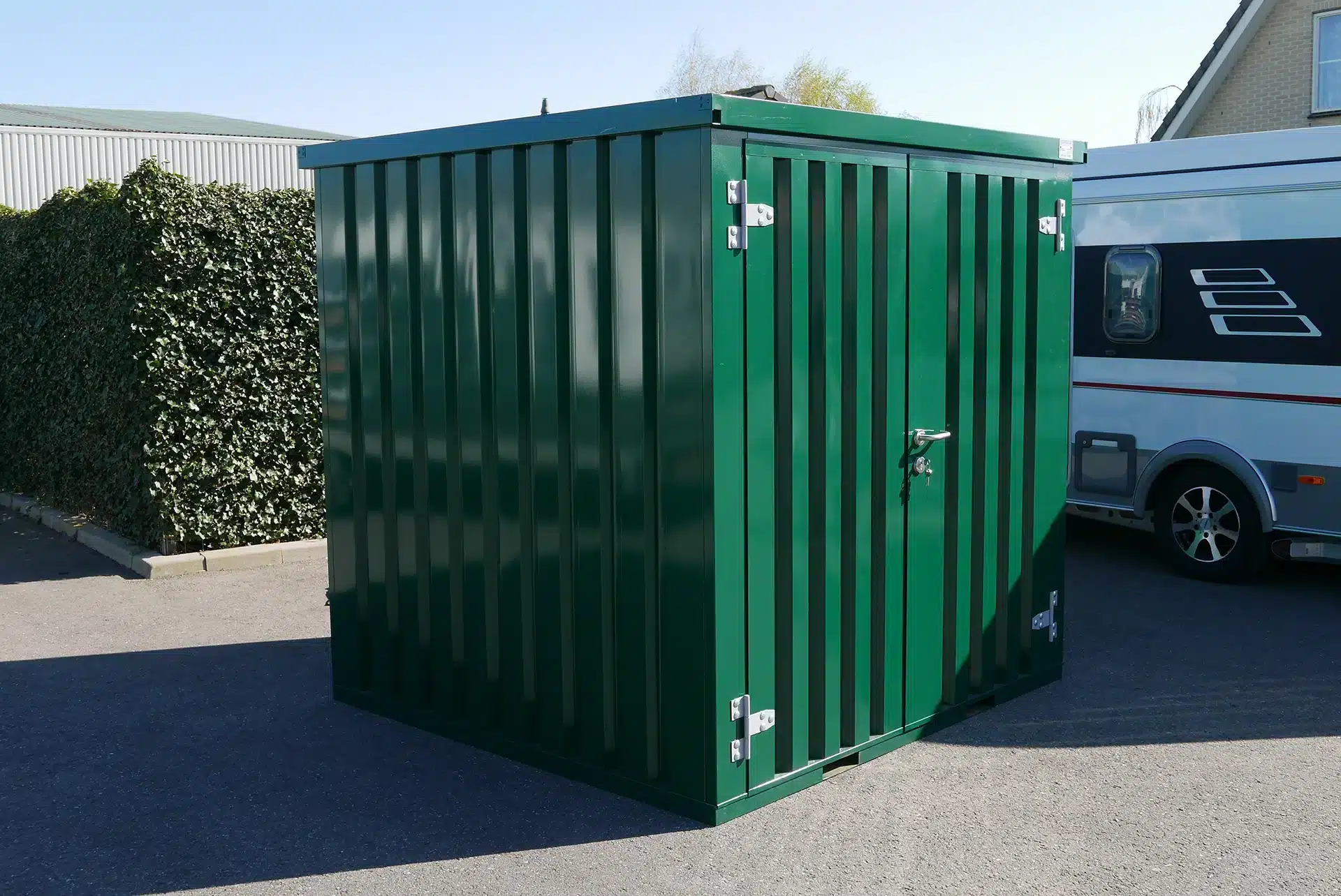 Een groene metalen opslagcontainer met een deur voorzien van zilverkleurige scharnieren en een slot. De container staat op een geasfalteerd terrein met een heg en een witte camper zichtbaar op de achtergrond. De lucht is helder en blauw, wat zorgt voor een zonnige belichting.