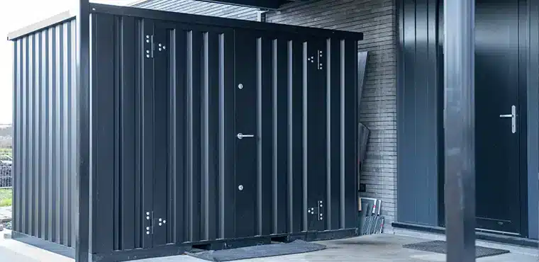 Een zwarte metalen opslagcontainer met dubbele deuren en zilverkleurige scharnieren, geplaatst onder een overkapping naast een grijs bakstenen gebouw. De container staat op een betonnen vloer, met enkele metalen platen en gereedschap zichtbaar op de achtergrond tegen de muur.
