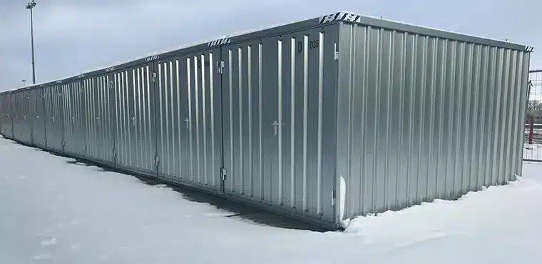 Een rij zilverkleurige metalen opslagcontainers met deuren en witte handgrepen, geplaatst op een met sneeuw bedekte ondergrond. De containers staan in een open gebied onder een heldere, bewolkte lucht, met lichte schaduwen zichtbaar op de sneeuw.
