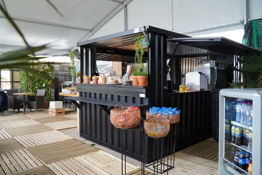 Zwarte container omgebouwd tot pop-up café met een toonbank, decoratieve planten en manden met snacks, geplaatst onder een tent met houten vlonders.