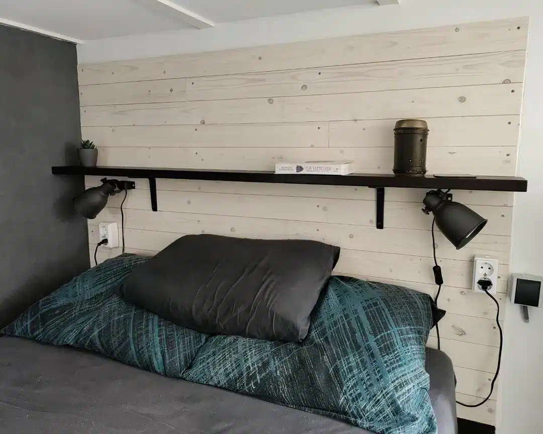Een slaapkamer met een bed dat bedekt is met een blauwgroene deken en een groot grijs kussen. De muur achter het bed is bekleed met lichte houten panelen. Een zwarte plank loopt horizontaal over de muur en bevat een klein plantje, een stapel boeken en een decoratieve metalen pot. Onder de plank hangen twee zwarte, verstelbare leeslampen die verbonden zijn met een witte stopcontactdoos aan beide kanten van het bed.