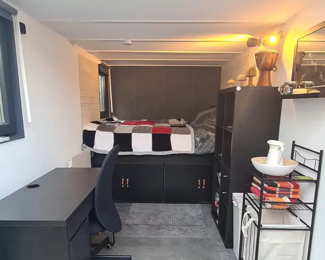 Een modern ingerichte kleine kamer met een bed tegen de achterwand, voorzien van een zwart-wit-rood dekbed en ingebouwde opbergladen eronder. Links staat een zwart bureau met een zwarte bureaustoel, terwijl rechts een zwarte open kast is geplaatst met decoratieve items zoals een beker en kleine voorwerpen. Naast de kast staat een metalen rek met handdoeken en een witte vaas. De kamer heeft grijze vloertegels en witte muren met een raam aan de linkerzijde dat zorgt voor natuurlijk licht.