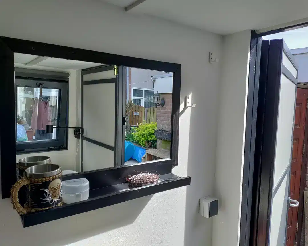Het interieur van een container met ramen met witte muren toont een groot rechthoekig raam met een zwart frame en een brede vensterbank. Op de vensterbank staan een kopje met een metalen thermoskan, een kleine witte container en een decoratieve borstel. Door het raam is de buitenruimte zichtbaar, inclusief een tuin met groen, een houten hek en een blauwe tafel. Rechts in beeld staat een open zwarte deur die naar buiten leidt.