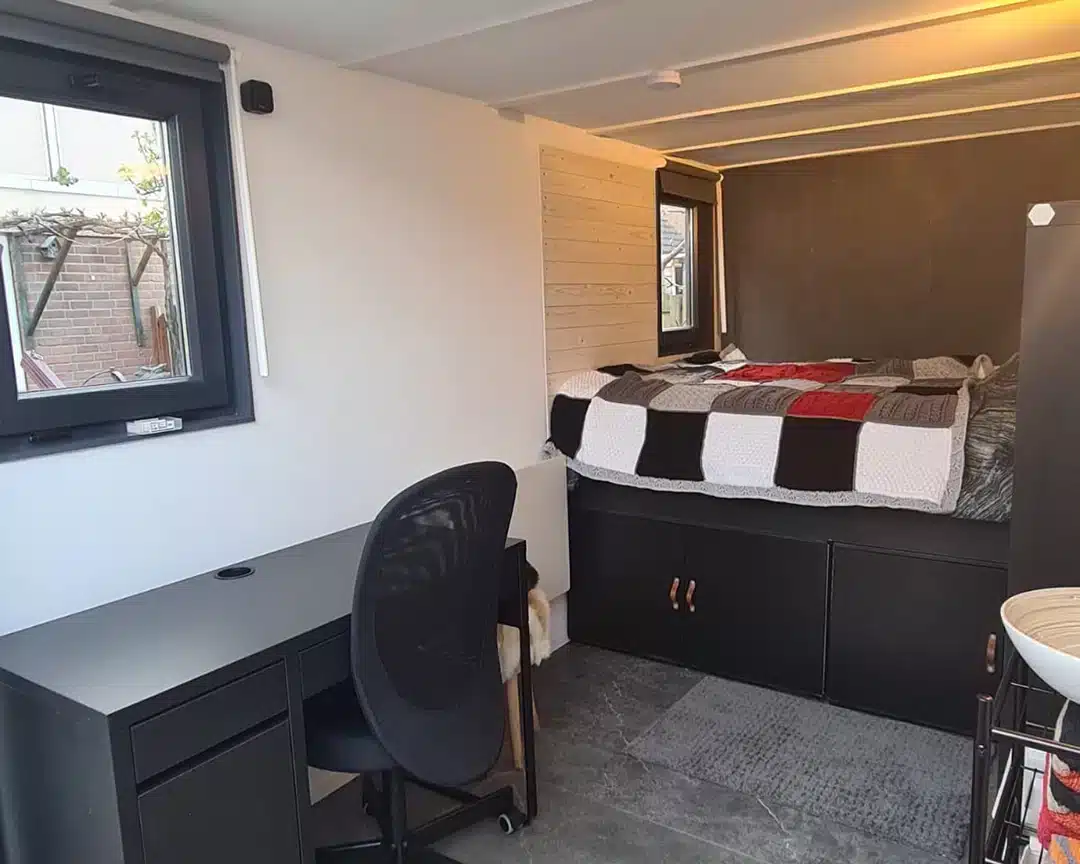 Een compacte slaapkamer met een ingebouwd bed dat voorzien is van een zwart-wit-rood blokkenpatroon dekbed. Onder het bed is er opbergruimte met zwarte kastdeuren. Links staat een zwart bureau met een bijpassende zwarte bureaustoel, geplaatst bij een raam dat uitkijkt op een tuin of binnenplaats. De vloer is afgewerkt met grijze tegels, en de muren zijn wit, met een gedeelte van lichte houten panelen.