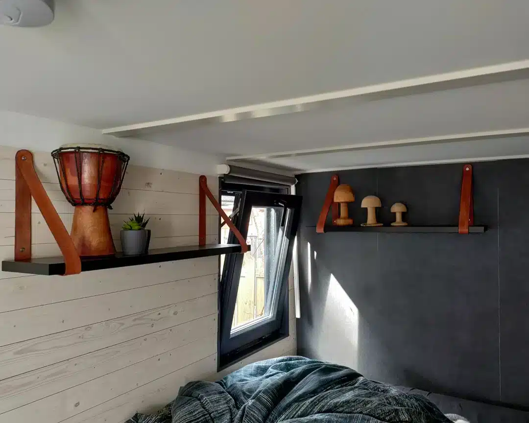 Een moderne slaapkamerhoek met een grijze wand en een raam dat natuurlijk licht binnenlaat. Boven het bed zijn twee zwarte planken met leren beugels gemonteerd. Op de linkerplank staat een decoratieve djembé-trommel en een kleine plant in een grijze pot. De rechterplank bevat houten decorstukken in de vorm van paddenstoelen. Het bed, gedeeltelijk zichtbaar, heeft een donkerkleurige deken.