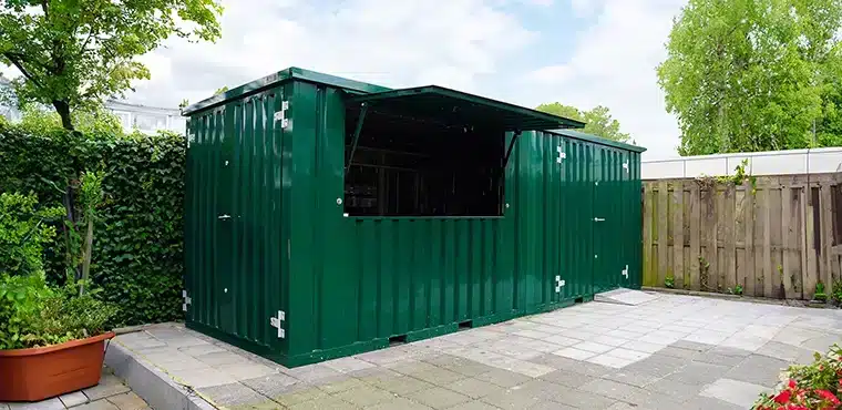 Een groene metalen container met een open luik dat als afdak dient, geplaatst op een betegeld terras. De container staat in een omheinde tuin, omgeven door groene struiken, bomen en een houten schutting. Het luik biedt een inkijk in de donkere binnenruimte van de container, die mogelijk dient als kiosk of opslagruimte