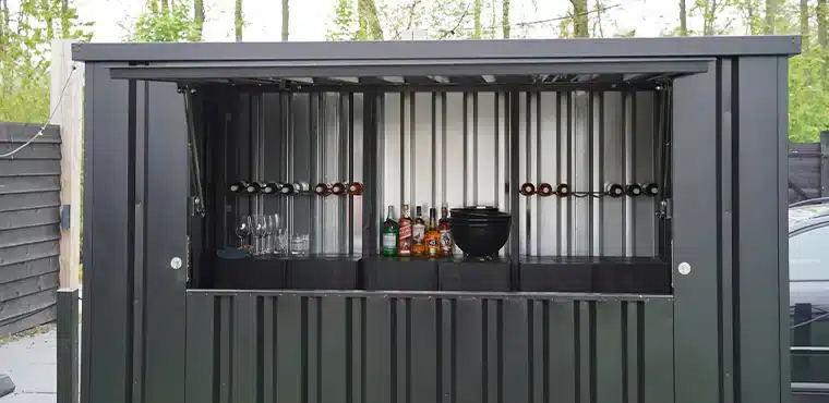 Een zwarte metalen container met een open luik dat dienst doet als bar. Binnenin zijn wijnflessen in rekken, glazen en verschillende flessen drank zichtbaar, samen met een zwarte kom op de toonbank. De achtergrond toont een houten schutting en enkele bomen, wat een buitenomgeving aanduidt.