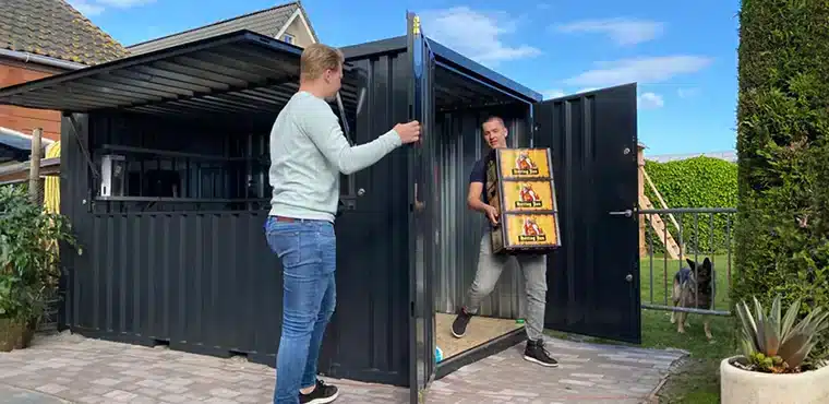 Een zwarte metalen container met een open deur en luik, geplaatst op een betegeld terras. Een man in een lichtblauwe trui houdt de deur open, terwijl een andere man een stapel kratten met drank draagt en naar buiten loopt. Op de achtergrond zijn een groene tuin, een schommel en een hond zichtbaar, wat een huiselijke omgeving aanduidt.