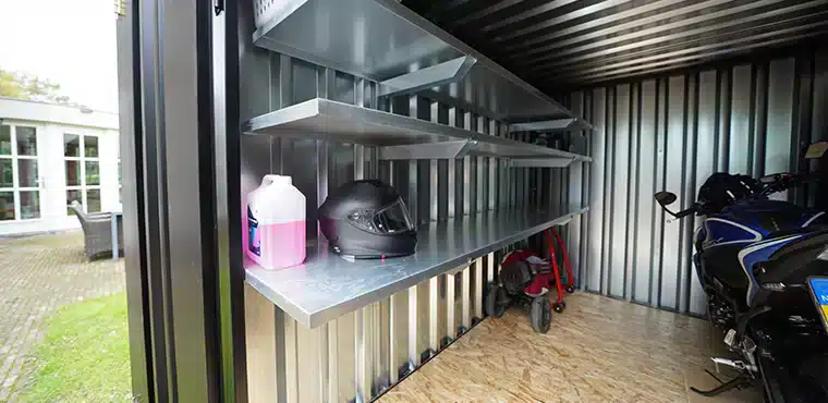Binnenkant van een metalen opslagcontainer met metalen rekken aan de linkerkant waarop een zwarte motorhelm en een roze jerrycan staan. Op de achtergrond staat een motorfiets en een step. De vloer is bedekt met houten platen en de wanden en het plafond zijn van gegalvaniseerd metaal. Door de open deur is er zicht op het grasveld buiten.