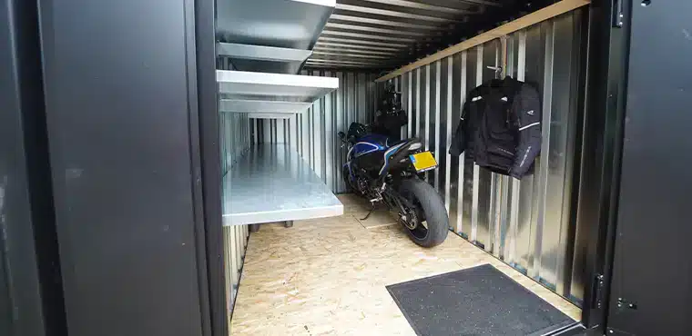 Binnenkant van een metalen opslagcontainer met aan de linkerzijde metalen rekken. Aan de rechterzijde staat een blauwe motorfiets met gele kentekenplaat en hangt een zwarte motorjas aan de wand. De vloer is bedekt met houten platen en er ligt een zwart matje bij de ingang. De wanden en het plafond zijn van gegalvaniseerd metaal.