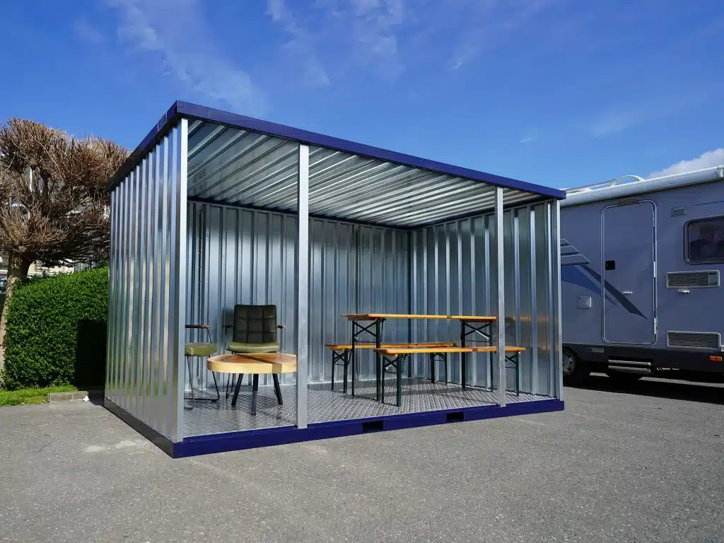 Een open metalen containerstructuur met een blauw frame en zilverkleurige panelen, ingericht als zitruimte. Binnenin staan een groene stoel en een kleine ronde houten tafel links, en een houten picknicktafel met bijpassende banken rechts. De container staat op een verhard oppervlak met een blauwe camper zichtbaar op de achtergrond en een boom aan de linkerzijde. De lucht is helder en blauw.