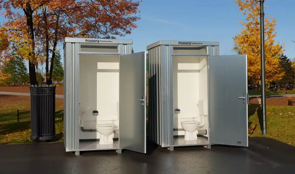 Twee mobiele sanitaire units met open deuren, elk met een toilet binnenin. De units zijn zilverkleurig en vervaardigd van metaal, geplaatst op een verharde ondergrond. De omgeving bestaat uit een parkachtig landschap met groen gras, bomen met herfstkleurige bladeren, een prullenbak, en straatlantaarns in de achtergrond. De lucht is helder en blauw.
