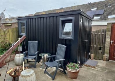 Zwarte demontabele container met ramen gebruikt als tuinhuis of buitenkantoor in een achtertuin met tuinmeubilair.
