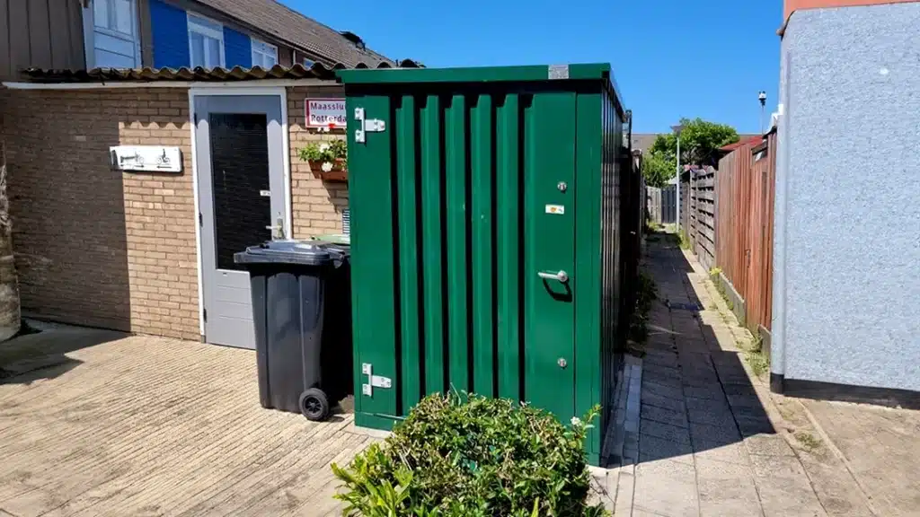 Een groene metalen opslagcontainer met een gesloten deur en zilverkleurige scharnieren, geplaatst op een betegeld pad naast een woning met een bakstenen gevel. Naast de container staat een zwarte afvalcontainer, en op de voorgrond is een kleine groene struik zichtbaar. De achtergrond toont een schutting en een heldere blauwe lucht.
