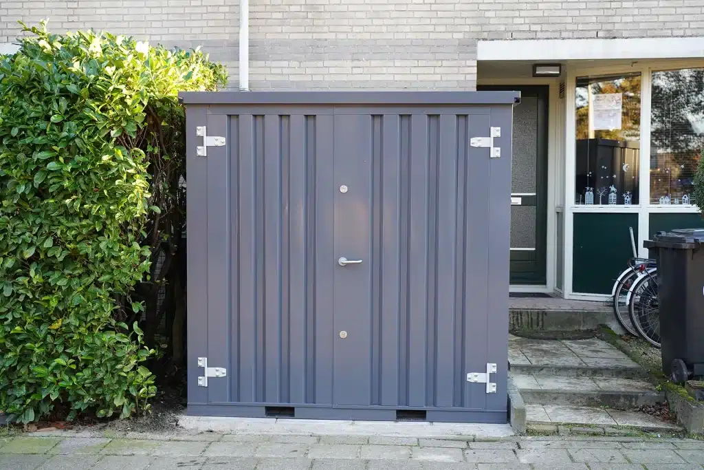 Een grijze metalen opslagcontainer met stevige dubbele deuren en zilverkleurige scharnieren, geplaatst op een betegelde ondergrond naast een groene heg. De container staat dicht bij de gevel van een bakstenen woonhuis, met een zwarte afvalcontainer, een fiets en een groene voordeur zichtbaar op de achtergrond.
