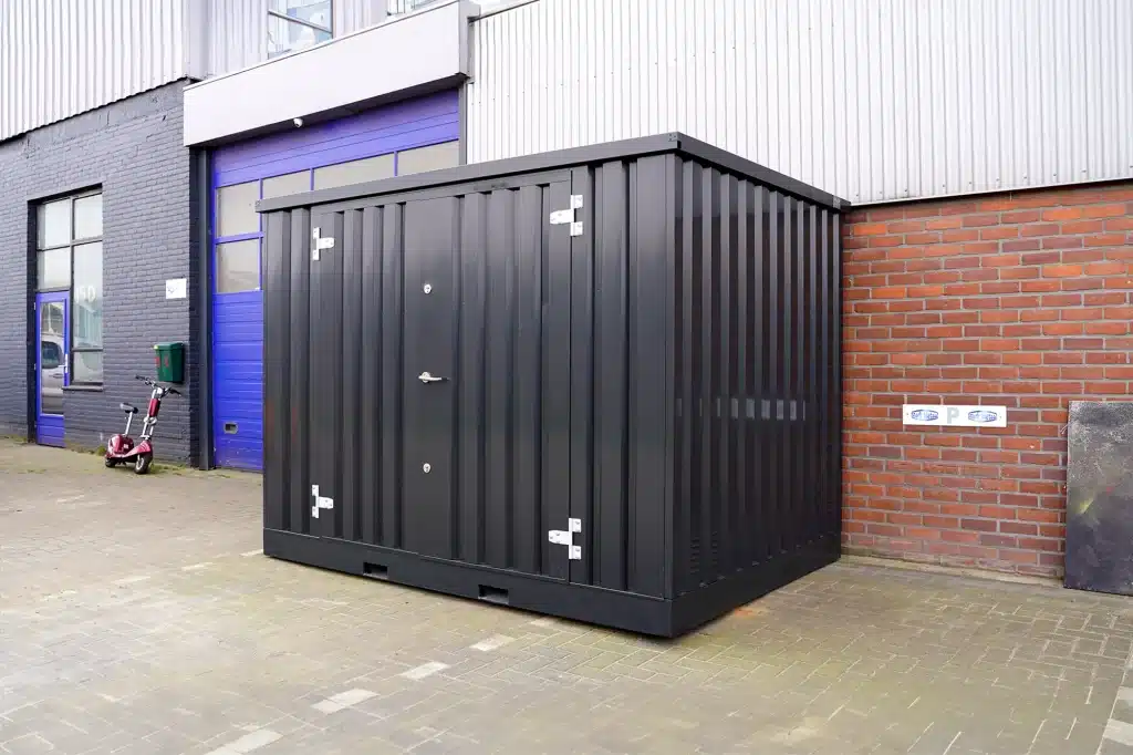 Een antraciete metalen opslagcontainer met gesloten dubbele deuren, geplaatst op een betegeld oppervlak naast een gebouw met een paarse deur en een bakstenen muur. De container is stevig gebouwd met metalen scharnieren en sloten. Op de achtergrond staat een deels zichtbare scooter tegen de muur.