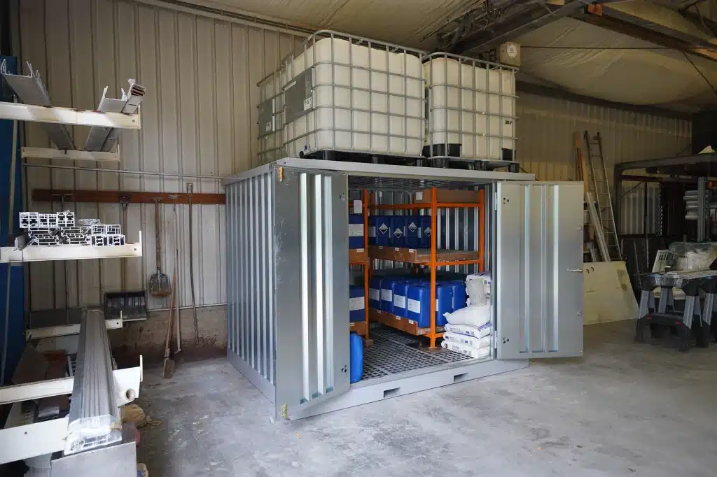 Een zilverkleurige metalen opslagcontainer in een loods met open deuren, waarin blauwe vaten en zakken met materialen op een oranje stellingkast zijn opgeslagen. Op het dak van de container staan twee grote witte watertanks. De loods heeft metalen wanden en een betonnen vloer, met enkele rekken en materialen zichtbaar aan de zijkanten.