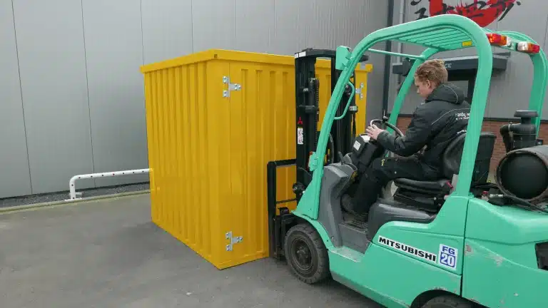 Een gele metalen opslagcontainer wordt verplaatst door een bestuurder op een mintgroene Mitsubishi vorkheftruck. De scène speelt zich af op een industrieterrein, met een grijze metalen gevel van een gebouw op de achtergrond en een witte buis langs de muur. De bestuurder draagt een zwarte jas en is gefocust op het bedienen van de heftruck.