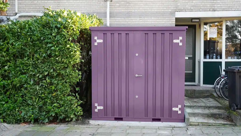 Een paarse metalen opslagcontainer met een deur voorzien van een zilverkleurig handvat en scharnieren. De container staat op een stoep naast een groene heg en is geplaatst tegen de gevel van een bakstenen woning. Op de achtergrond zijn een voordeur, een raam en een deel van een fiets zichtbaar.