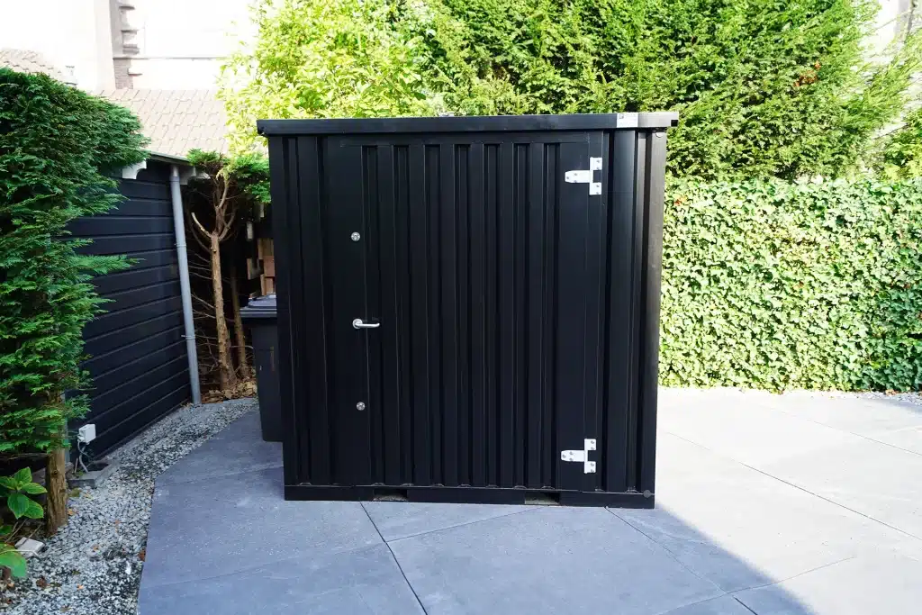 Een zwarte metalen 8ft opslagcontainer met een stevige deur, geplaatst op een betegelde ondergrond in een tuin. De container wordt omringd door groene heggen en struiken, met een deel van een houten schuurtje zichtbaar aan de linkerkant. Het gebied is goed verlicht door zonlicht.