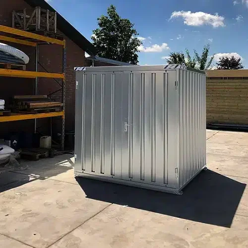 Een metalen 8ft opslagcontainer geplaatst op een zonnig buitenterrein. De container heeft een afsluitbare deur aan de voorkant en staat naast een rek met bouwmaterialen. Op de achtergrond zijn een houten schutting, een bakstenen gebouw en enkele bomen zichtbaar onder een heldere blauwe lucht.